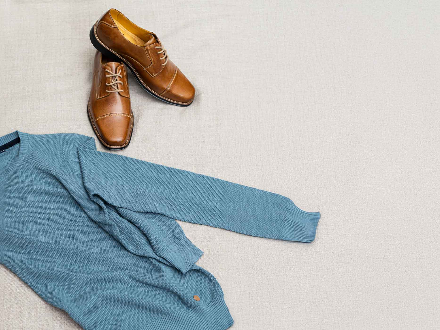 Ein blauer Pullover liegt gefaltet auf einem wei&szlig;en Teppich, daneben liegt ein Paar braune Herrenschuhe.