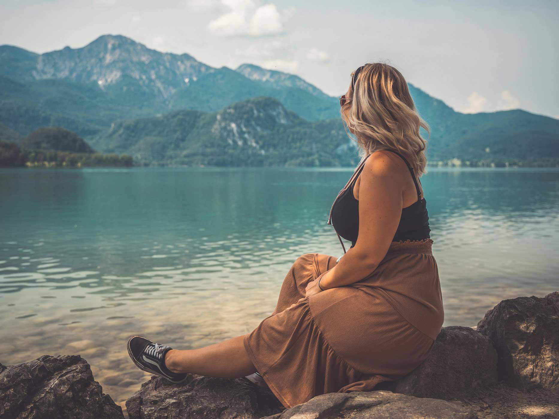 Curvy Frau sitzt auf Steinen vor dem Meer und trägt ein schwarzes Oberteil und einen leichten, langen braunen Rock